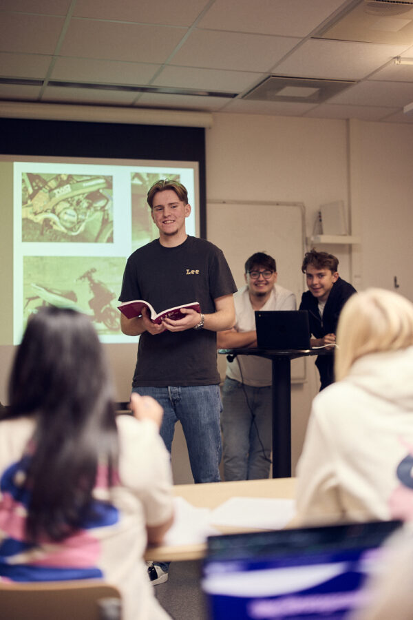 Elev står längst fram i klassrummet och håller en presentation för klassen medan de andra eleverna lyssnar.