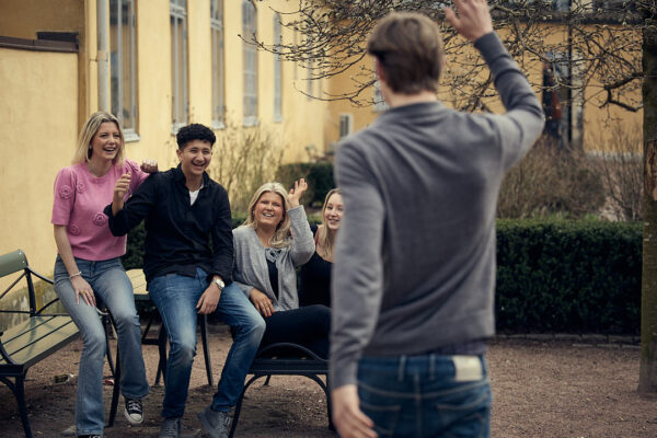 Fyra gymnasieelever sitter utomhus på en bänk och skrattar medan de vinkar glatt till en elev som närmar sig dem.