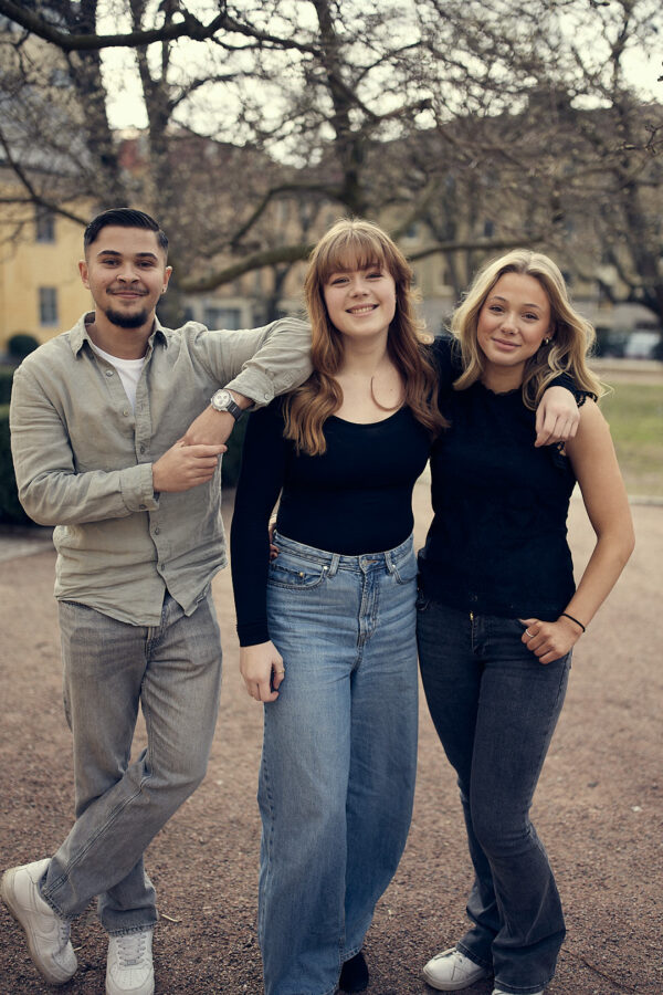 Tre gymnasieelever står tillsammans utomhus och poserar glatt för kameran, med armarna runt varandra i en avslappnad miljö.