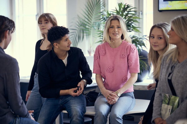 Sex gymnasieelever står och sitter tillsammans i ett klassrum med stora fönster. Fokus är på en tjej i rosa tröja som tittar mot kameran.